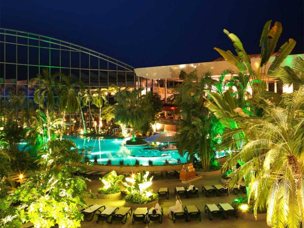 Abendaufnahme der Therme Erding mit Palmen, beleuchtetem Thermalbecken und tropischer Atmosphäre – Blick auf die Vital Oase und den Thermenbereich.