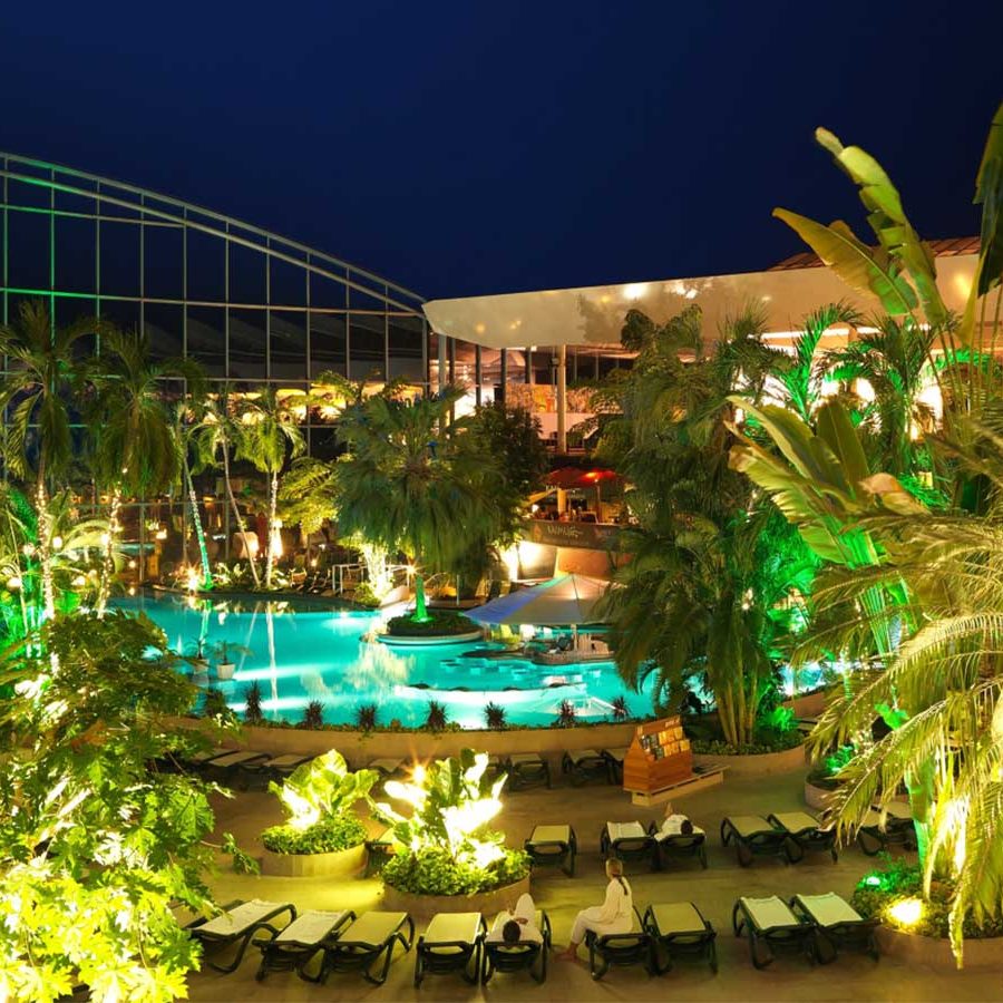 Abendaufnahme der Therme Erding mit Palmen, beleuchtetem Thermalbecken und tropischer Atmosphäre – Blick auf die Vital Oase und den Thermenbereich.
